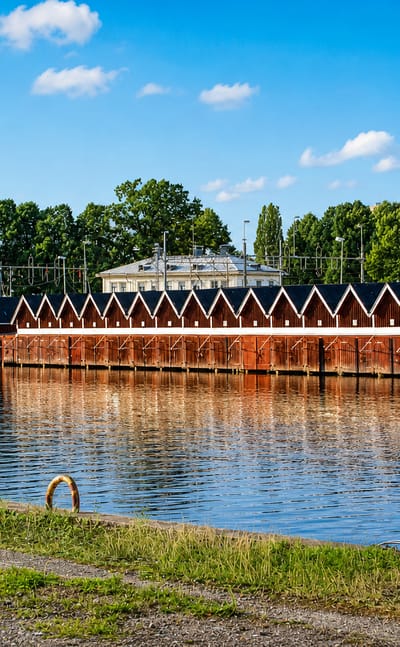 Row of red wooden boathouses reflected in a calm canal under blue sky at Köping, Sweden