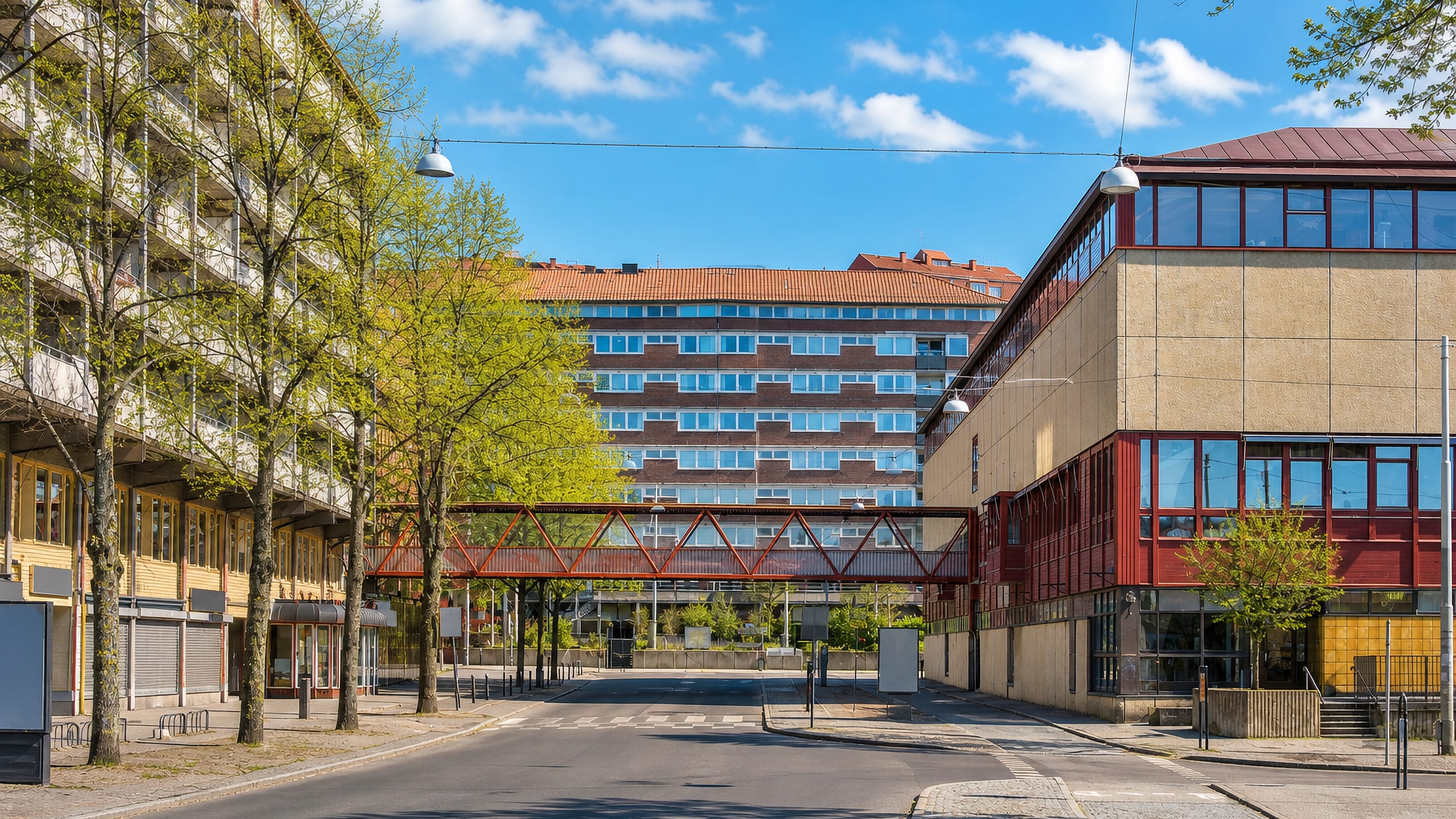 Red steel walkway bridge linking modernist blocks above a quiet street in Landala, central Gothenburg.