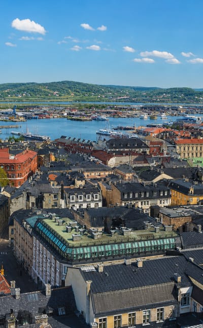 Aerial daytime view across the rooftops of Nordstaden, Sweden toward a harbour and wooded hills under a clear blue sky.