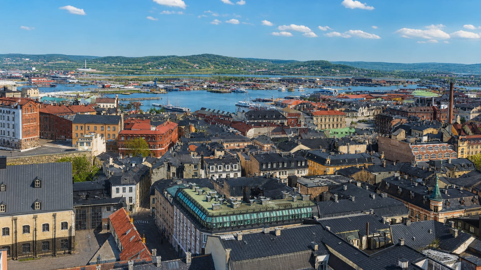 Aerial daytime view across the rooftops of Nordstaden, Sweden toward a harbour and wooded hills under a clear blue sky.