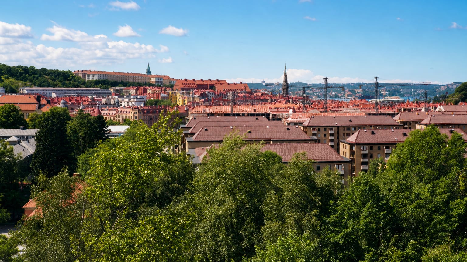 Hilltop view across the red rooftops of Olivedal, Gothenburg, with green treetops, a church spire and a distant bridge.