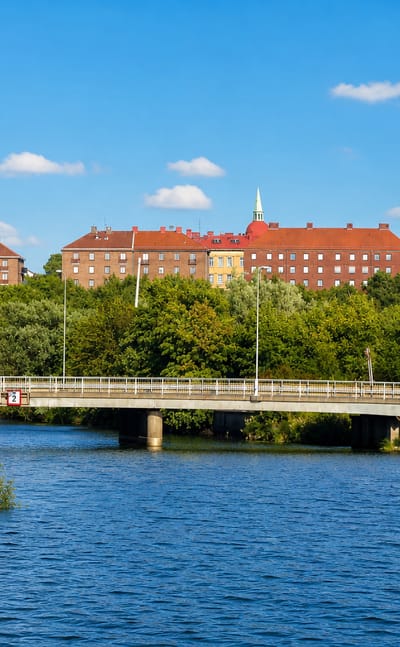 Red-brick apartment blocks above tree-lined water and a road bridge in Olskroken, Gothenburg, under a blue summer sky