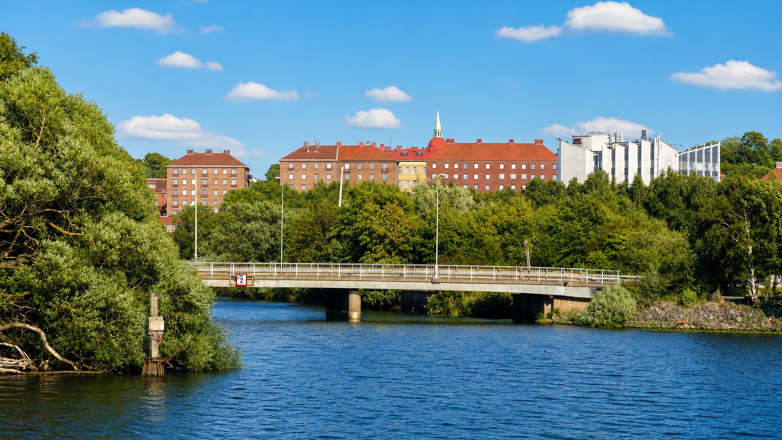 Red-brick apartment blocks above tree-lined water and a road bridge in Olskroken, Gothenburg, under a blue summer sky