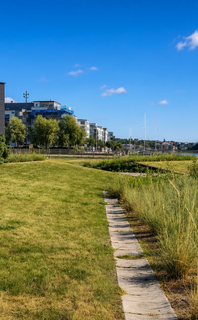 Modern waterfront apartment blocks beside a grassy harbourside park under blue sky in Sannegården, Gothenburg, Sweden.