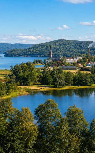Elevated summer view over Mellerud, Sweden, with a foreground lake, forested hills and the lakeside town's church spire.