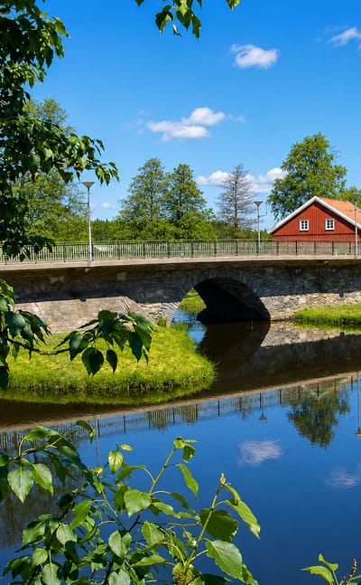 Stone arch bridge over the calm Nossan river in Nossebro, Sweden, with a red cottage and summer trees reflected in the water.