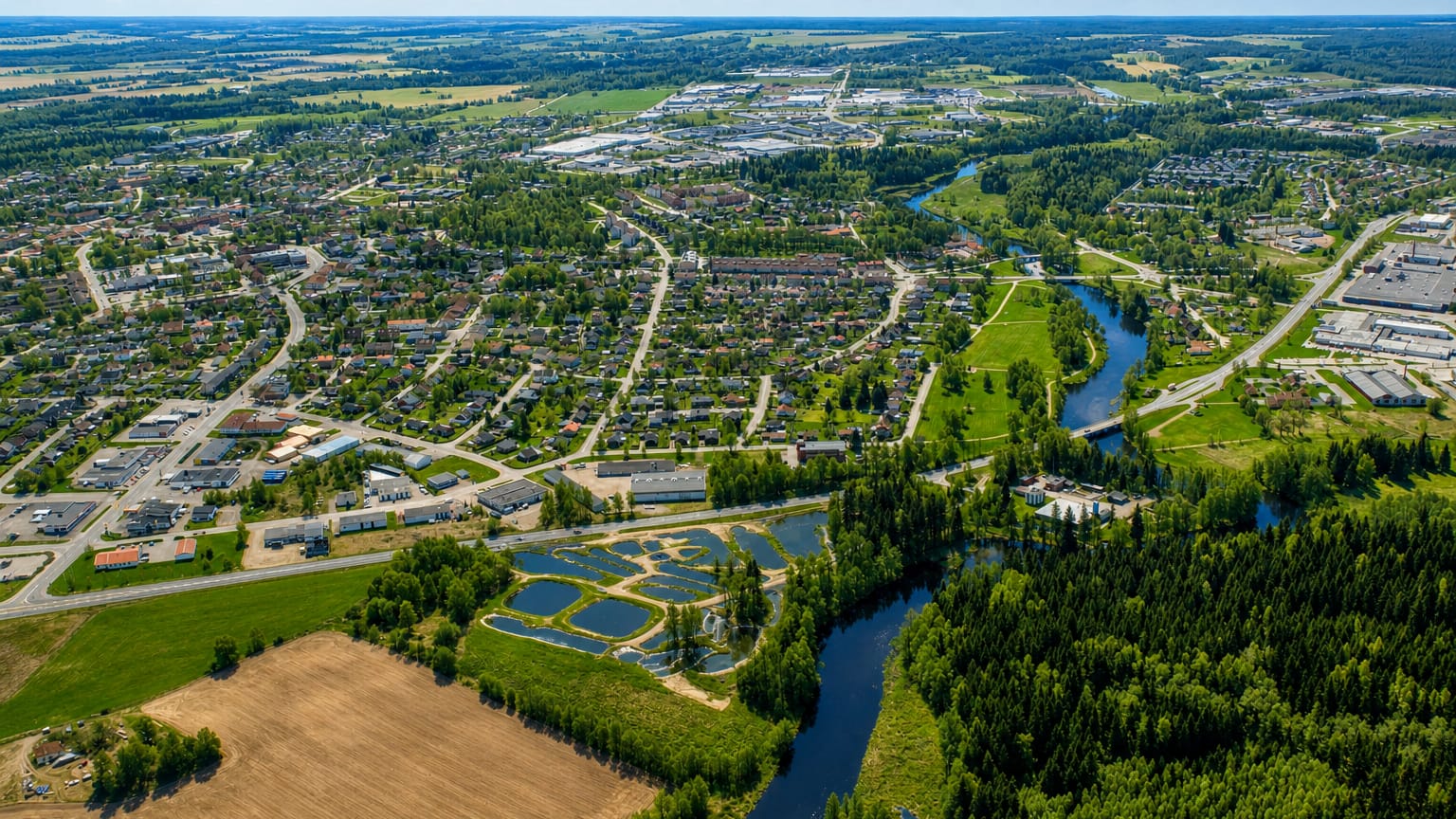 Aerial view of Tibro, Sweden, a small town with a winding river, residential streets and surrounding forest under a blue summer sky.
