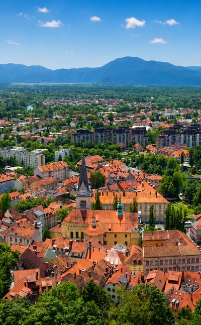 Aerial view of Ljubljana's red-roofed old town and church tower beside the river, framed by wooded hills and mountains.