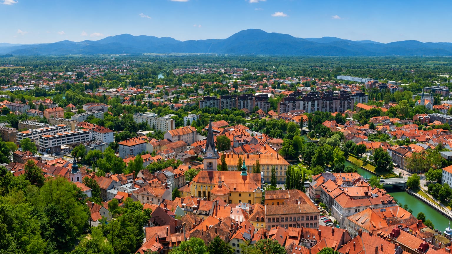 Aerial view of Ljubljana's red-roofed old town and church tower beside the river, framed by wooded hills and mountains.