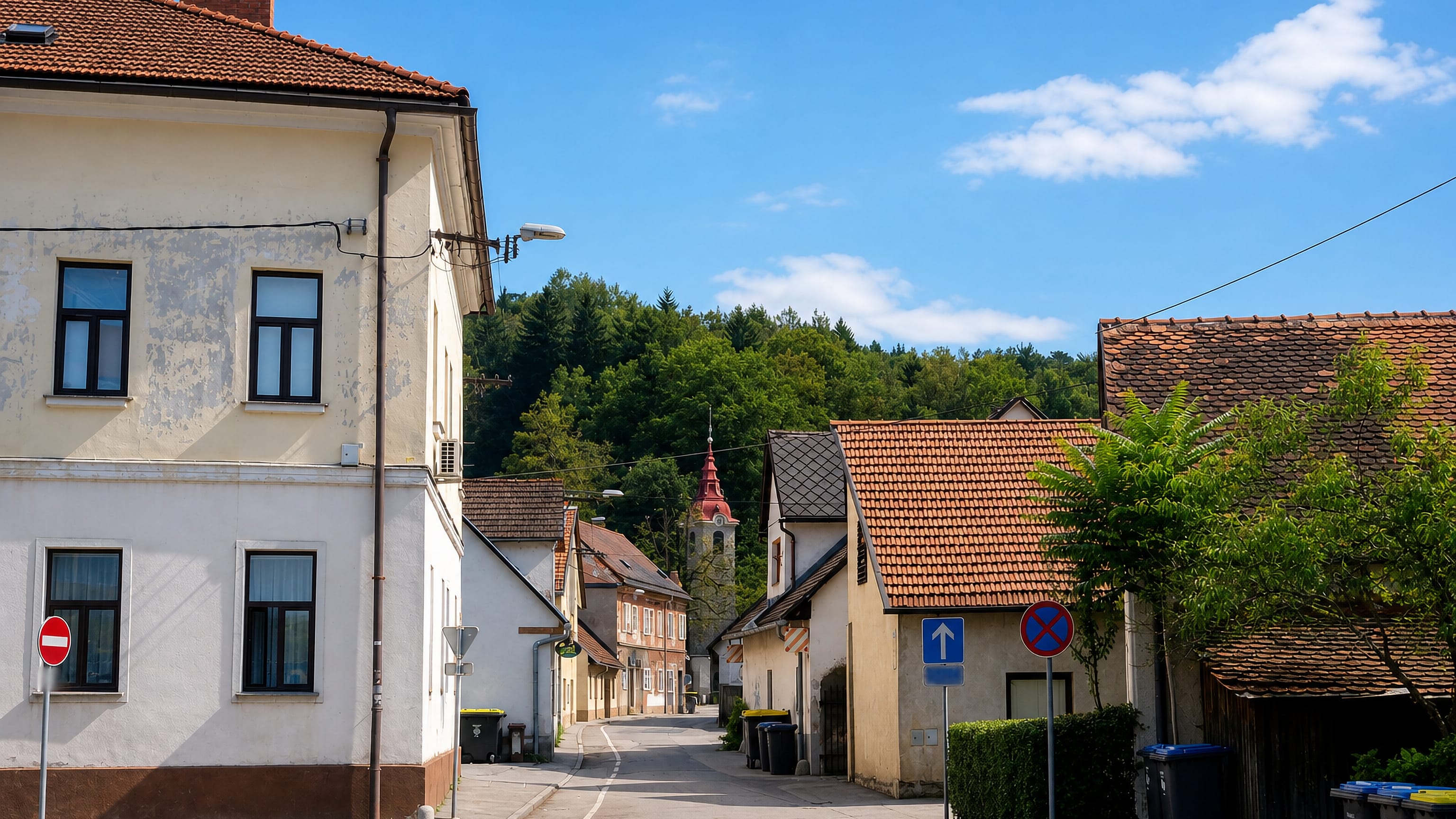 Old village street with red-tiled roofs and a church tower below wooded hills in Spodnja Šiška, Ljubljana.