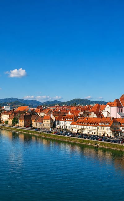 Maribor's old town and red-roofed Lent waterfront reflected in the Drava River below green hills, Slovenia.