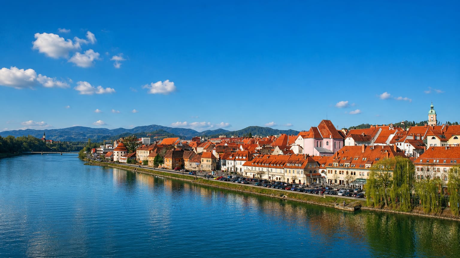 Maribor's old town and red-roofed Lent waterfront reflected in the Drava River below green hills, Slovenia.