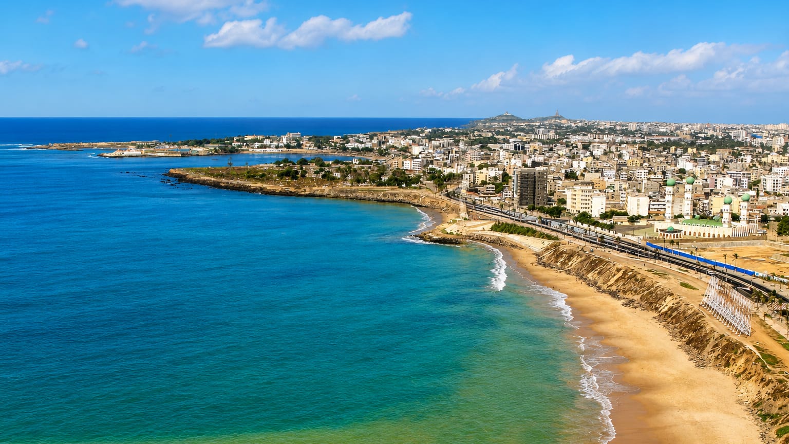 Dakar's Atlantic coastline in daylight, a curving beach and corniche below the city's rooftops and a mosque.