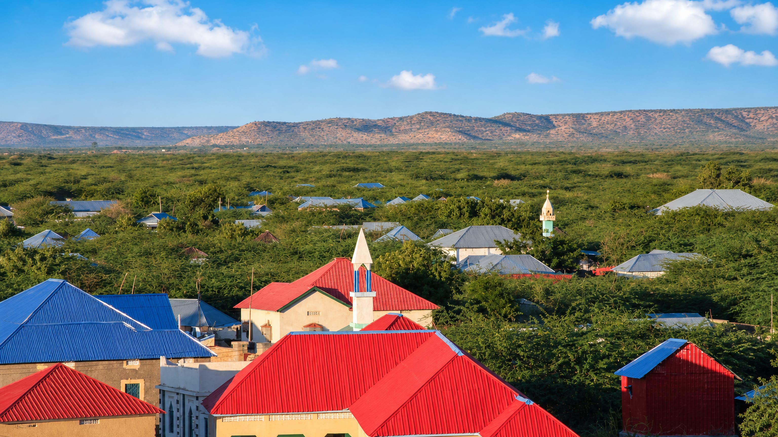 Red and blue tin roofs and a slender minaret among acacia scrub at Beledweyne, Somalia, with hills beyond