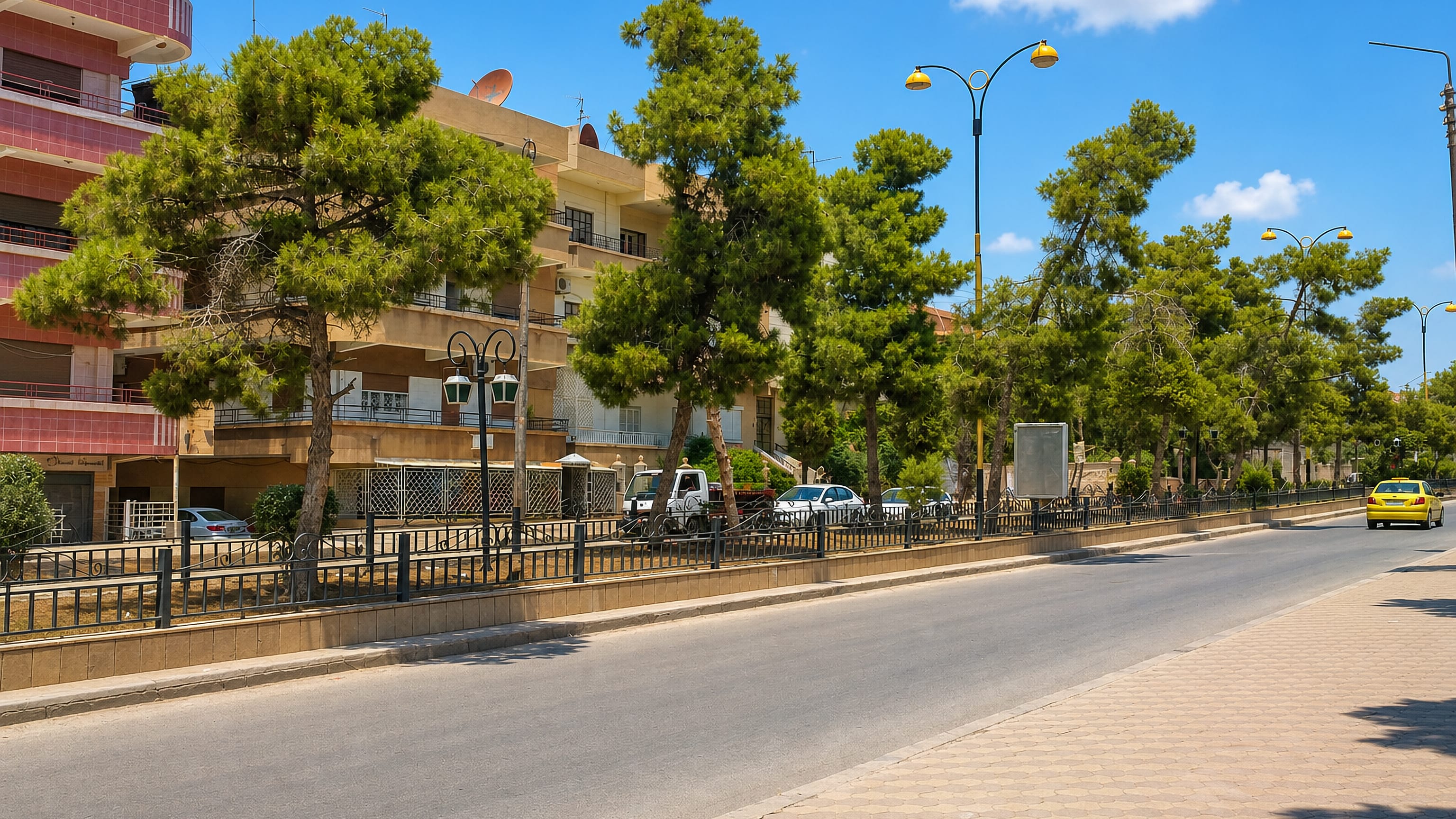 A pine-lined boulevard in Al-Qamishli, Syria, with mid-rise apartment blocks and a yellow taxi.
