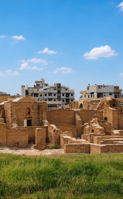 Ancient brick ruins beside modern apartment blocks in Ar-Raqqah under a bright sky.