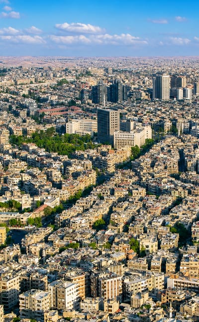 Dense aerial townscape of Damascus with sprawling low-rise buildings, scattered minarets and mosque domes, and hills beyond