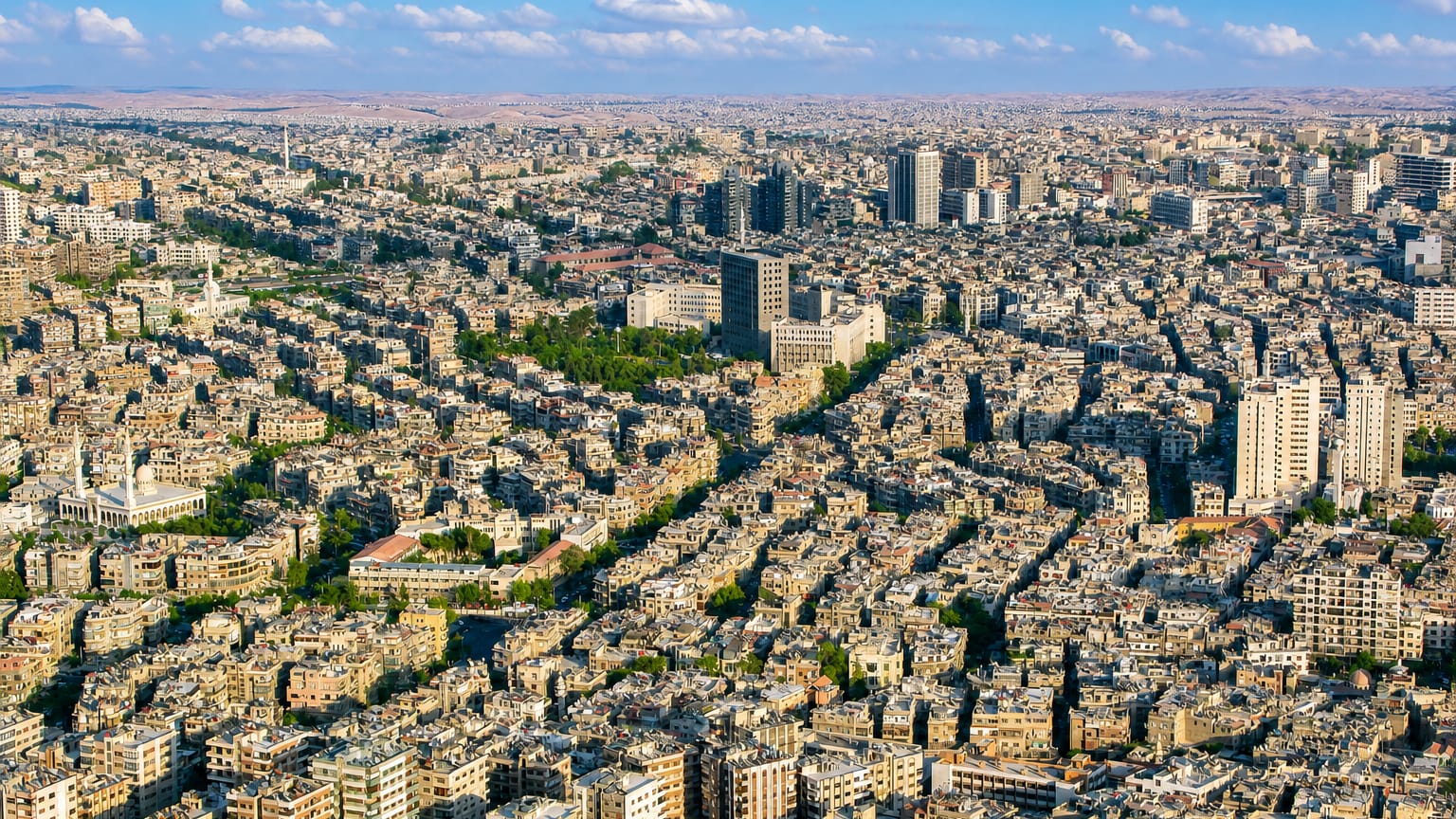 Dense aerial townscape of Damascus with sprawling low-rise buildings, scattered minarets and mosque domes, and hills beyond