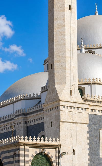 White-domed Khalid ibn al-Walid Mosque with its tall minaret under a blue sky in Homs.