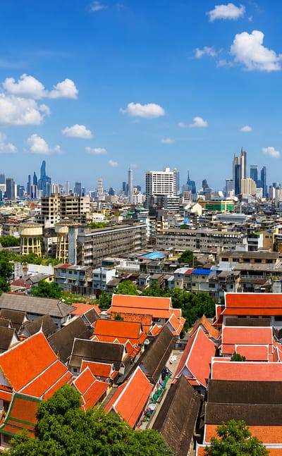 Bangkok's orange-tiled temple roofs in the foreground with the modern high-rise skyline beyond under blue sky.