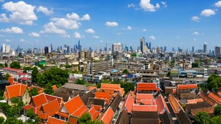 Bangkok's orange-tiled temple roofs in the foreground with the modern high-rise skyline beyond under blue sky.