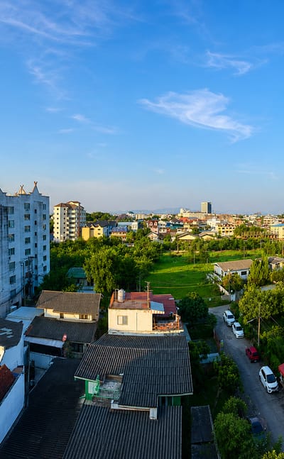 Chiang Mai rooftops with a golden temple spire, lush trees and distant mountains in warm morning light.