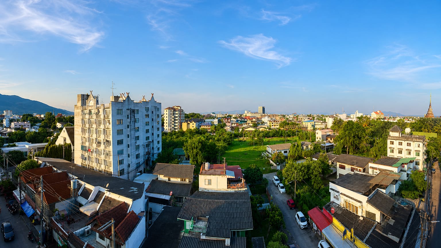 Chiang Mai rooftops with a golden temple spire, lush trees and distant mountains in warm morning light.
