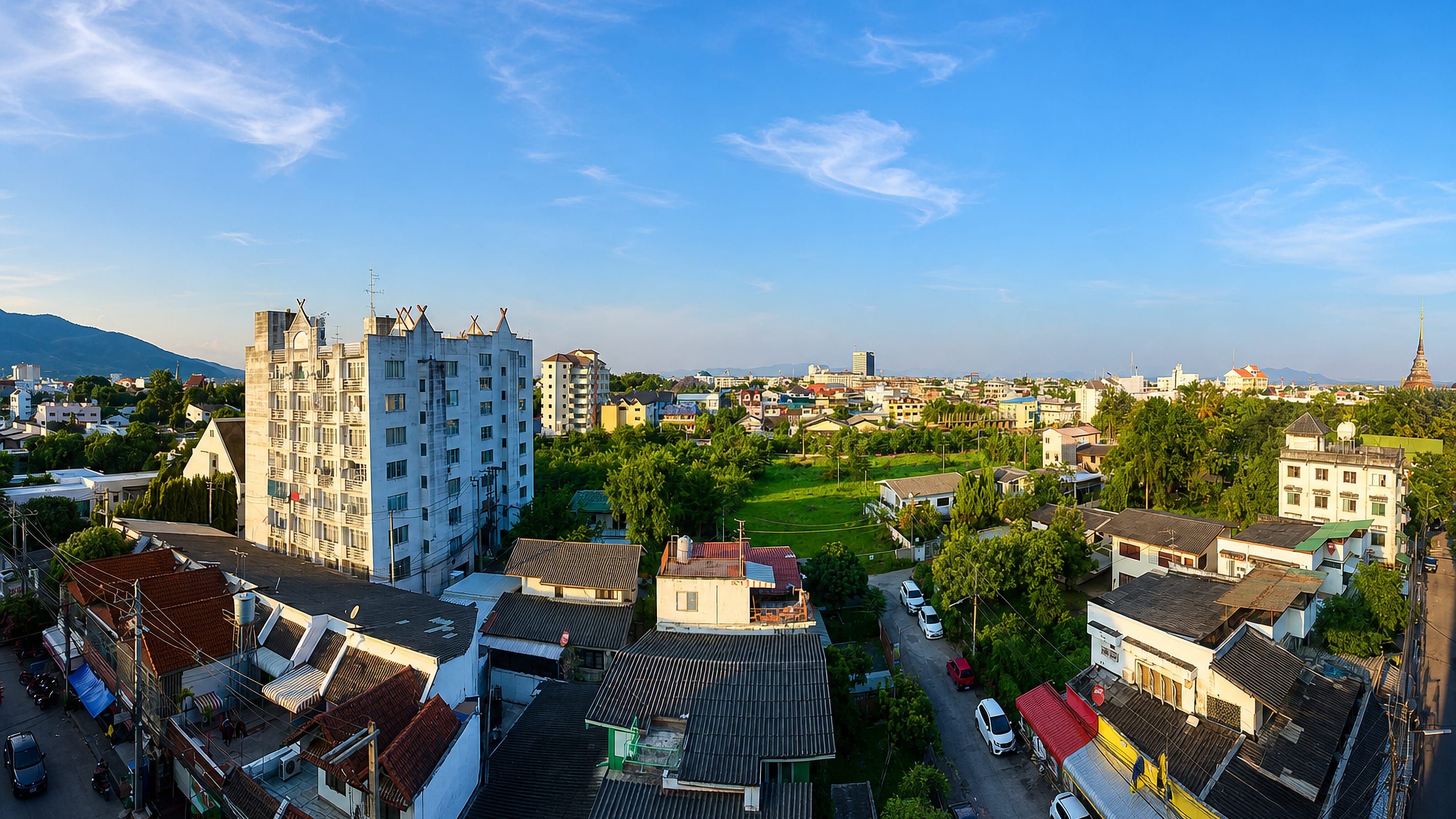 Chiang Mai rooftops with a golden temple spire, lush trees and distant mountains in warm morning light.