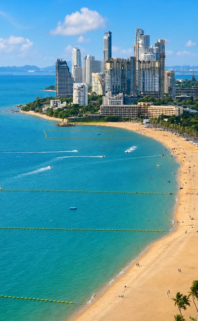 Pattaya Beach in daylight, a curving sandy bay lined with high-rise hotels and palms, boats on turquoise water.