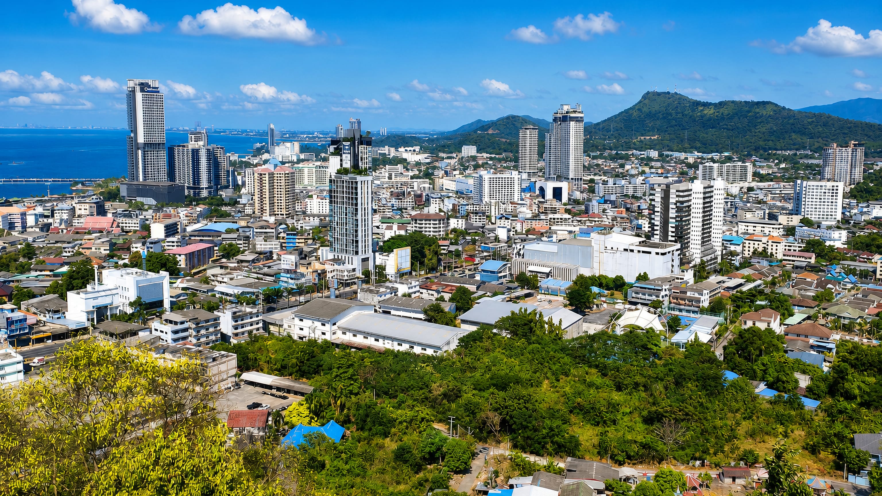 Hillside view over Si Racha's towers and rooftops toward the Gulf of Thailand and green hills.