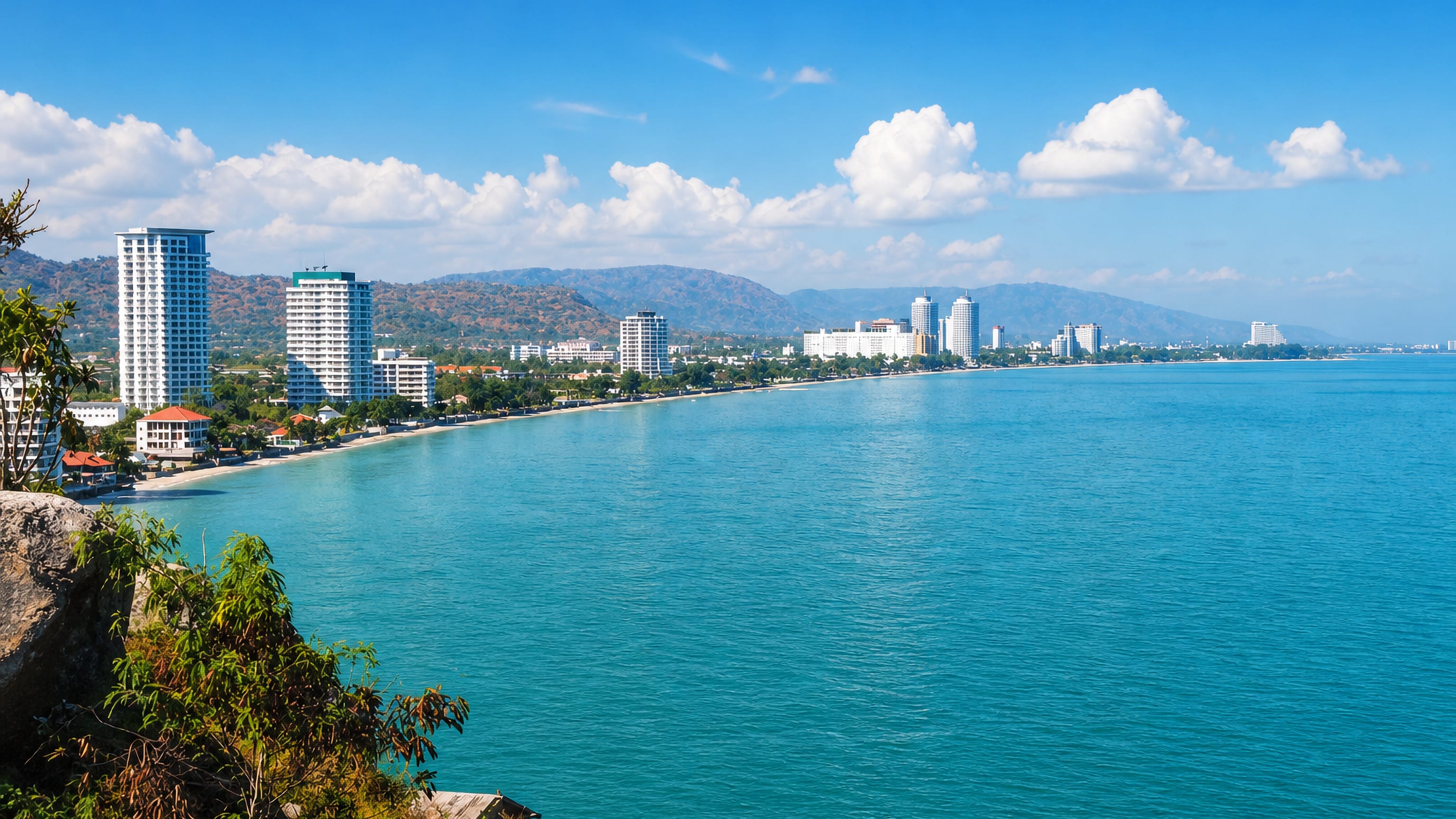The curving beachfront of Hua Hin, Thailand, lined with hotel towers below forested hills.