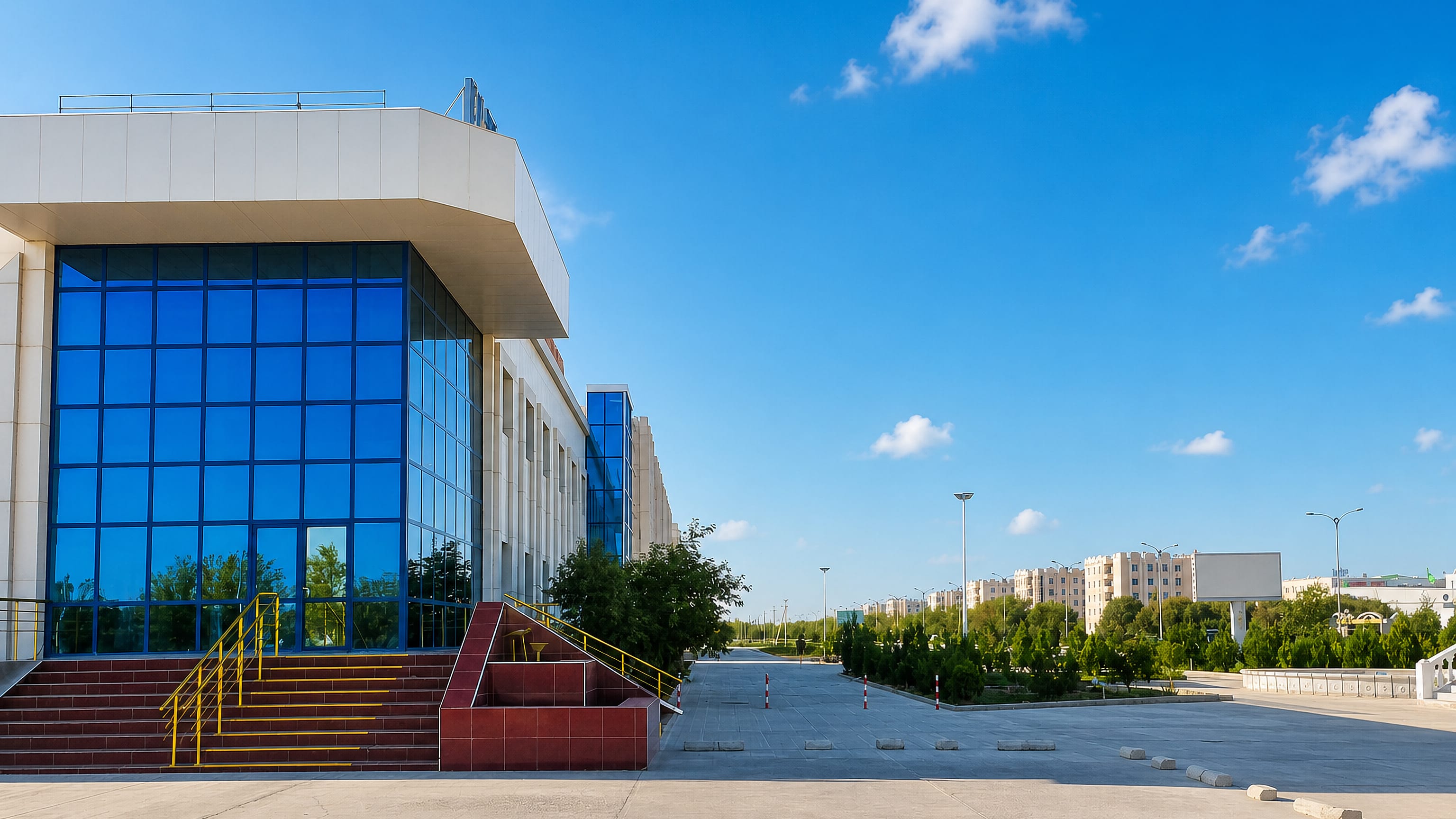 Modern blue-glass civic building beside a wide paved plaza under clear skies in Dasoguz, Turkmenistan.