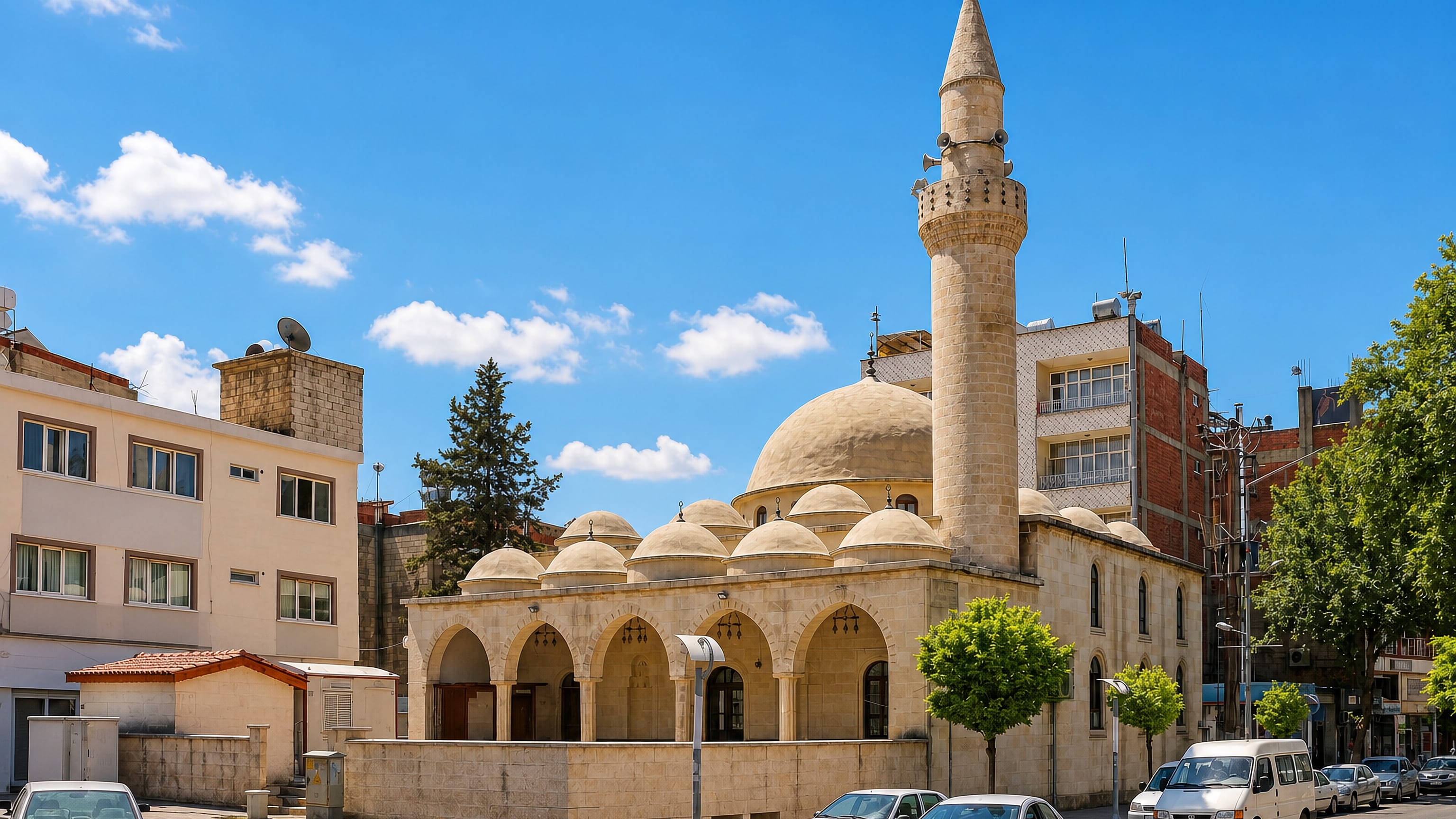 A domed Ottoman-style mosque and stone minaret on a sunlit street corner in Adıyaman, Turkey.