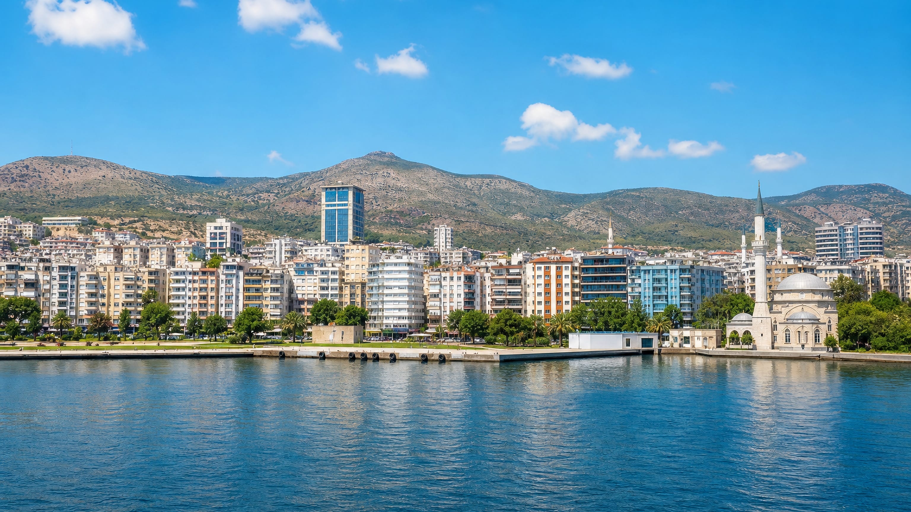 Waterfront apartment blocks and a domed mosque along Izmir Bay at Cigli, with dry hills behind.