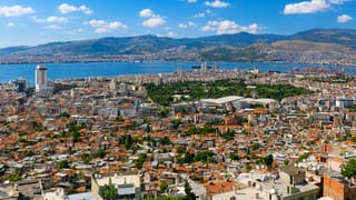 Aerial view of İzmir's red-roofed cityscape along its bay with mountains and port cranes beyond