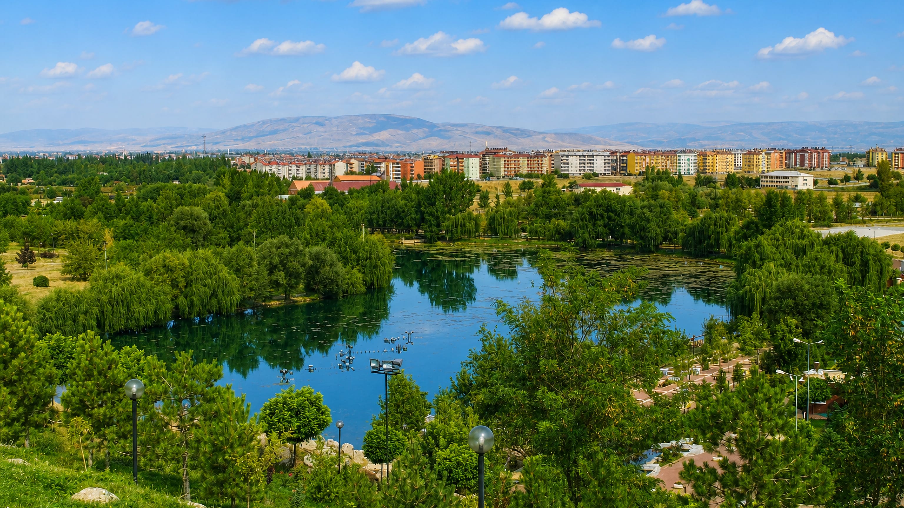 Park lake fringed with trees in front of Elbistan's apartment blocks and the mountains of Kahramanmaraş.