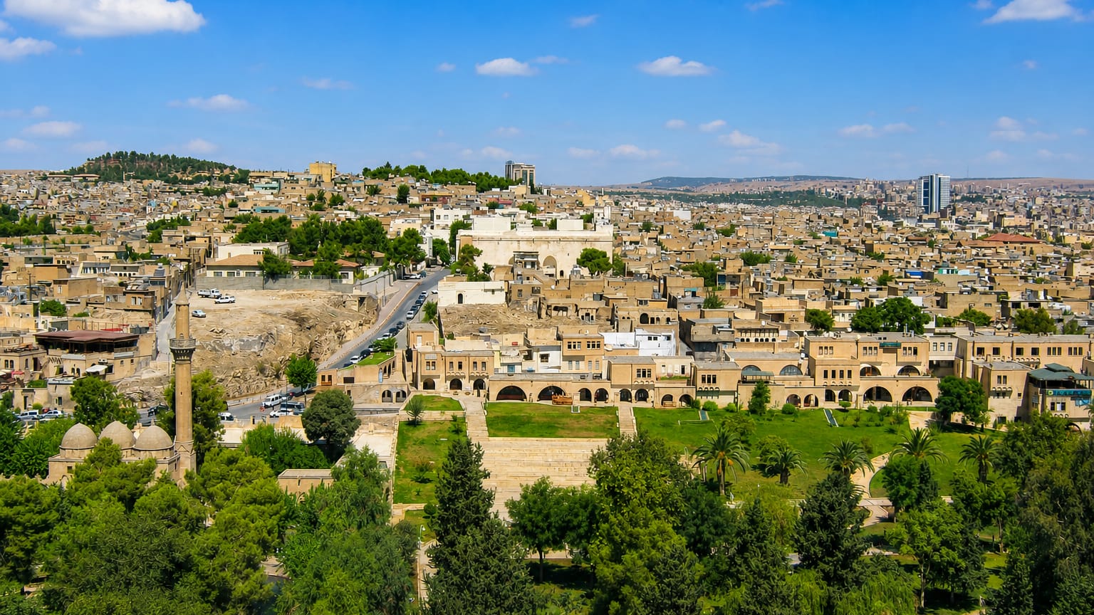 Sanliurfa's tan stone old town spreading over hills, with a minaret and the green Golbasi gardens below
