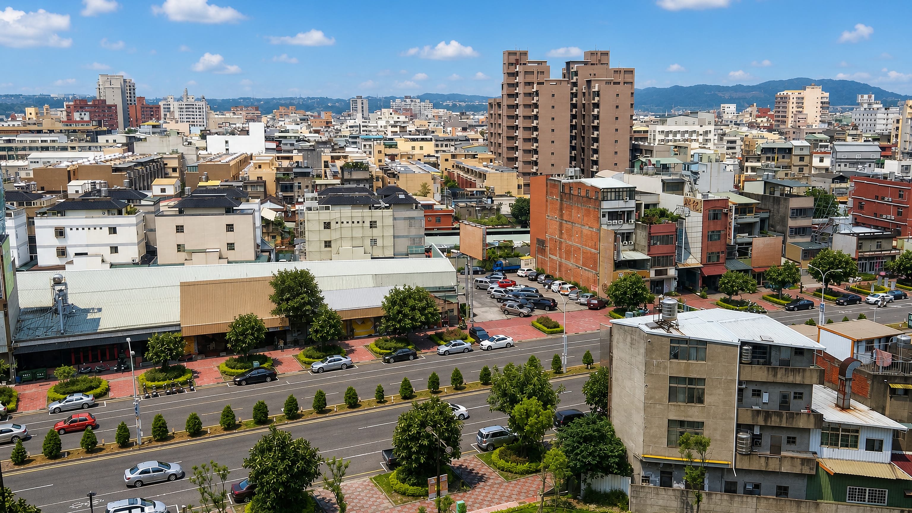 Elevated view over Toufen, Taiwan, with low-rise blocks, a tree-lined boulevard and green hills beyond.