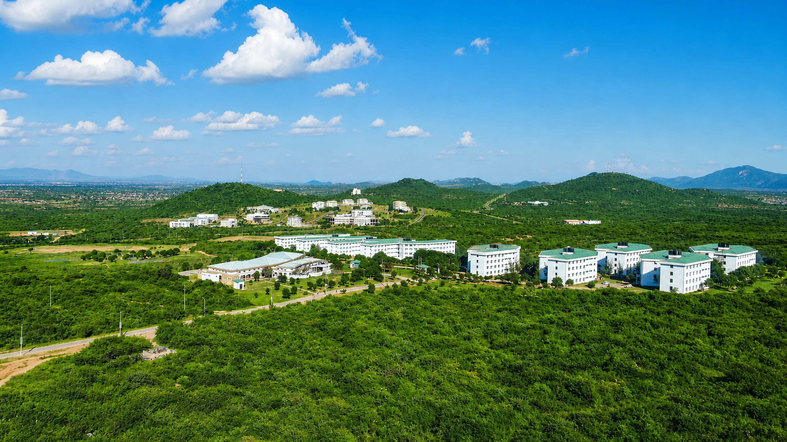 Hillside campus buildings with green roofs above scrubland on the edge of Dodoma, Tanzania, under a blue sky.