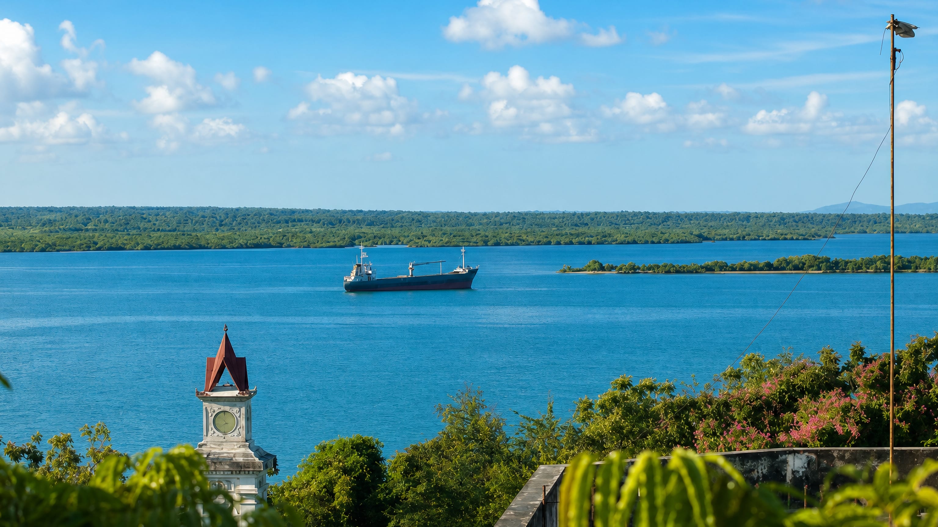 Cargo ship in Tanga bay, Tanzania, seen past a red-roofed colonial clock tower and mangrove shore.