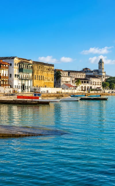 Zanzibar Stone Town seafront, Tanzania, with historic buildings, a clock tower and boats on turquoise water.