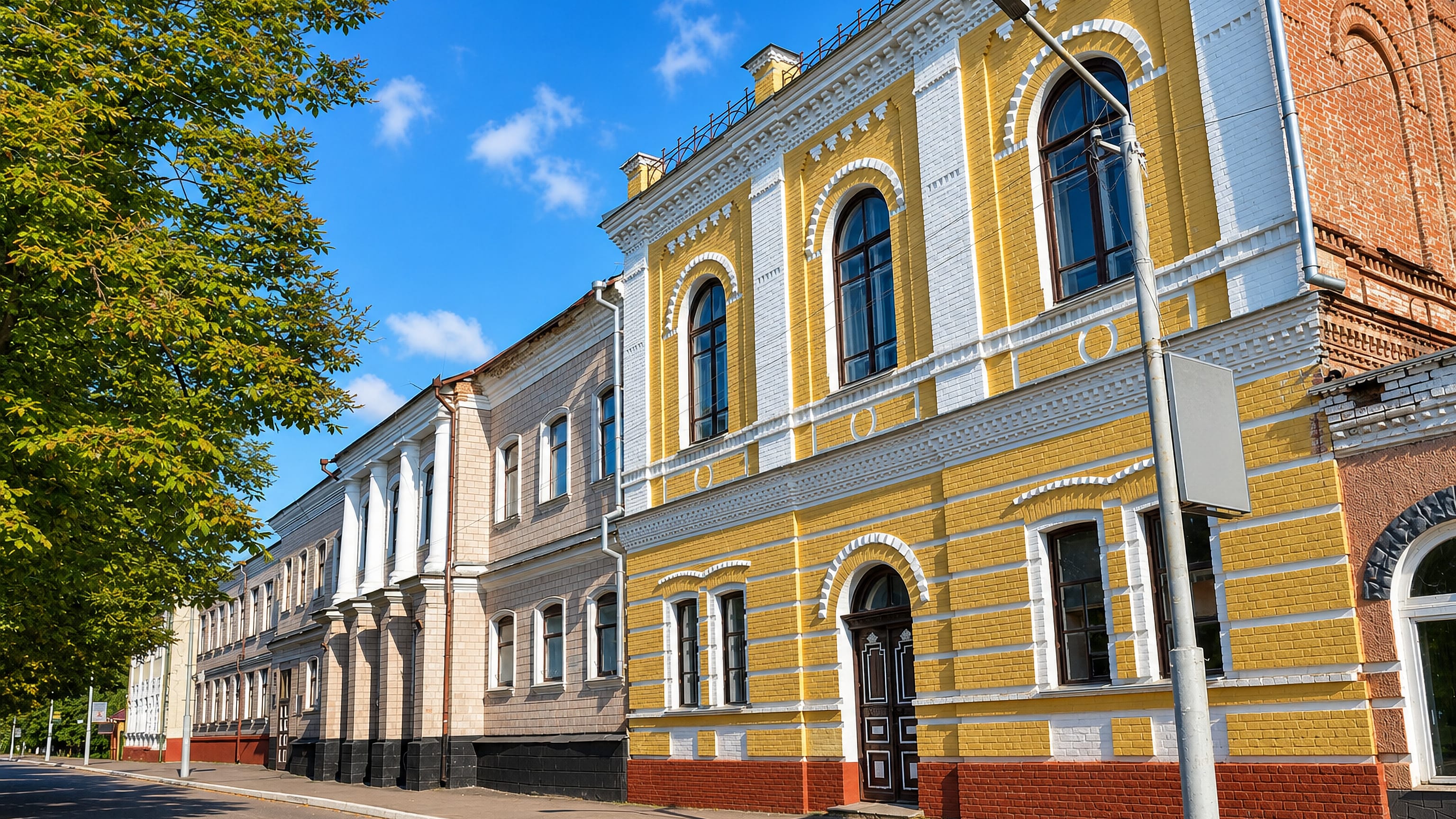 Sunlit 19th-century yellow and white brick facades lining a quiet tree-shaded street in Nizhyn, Ukraine.