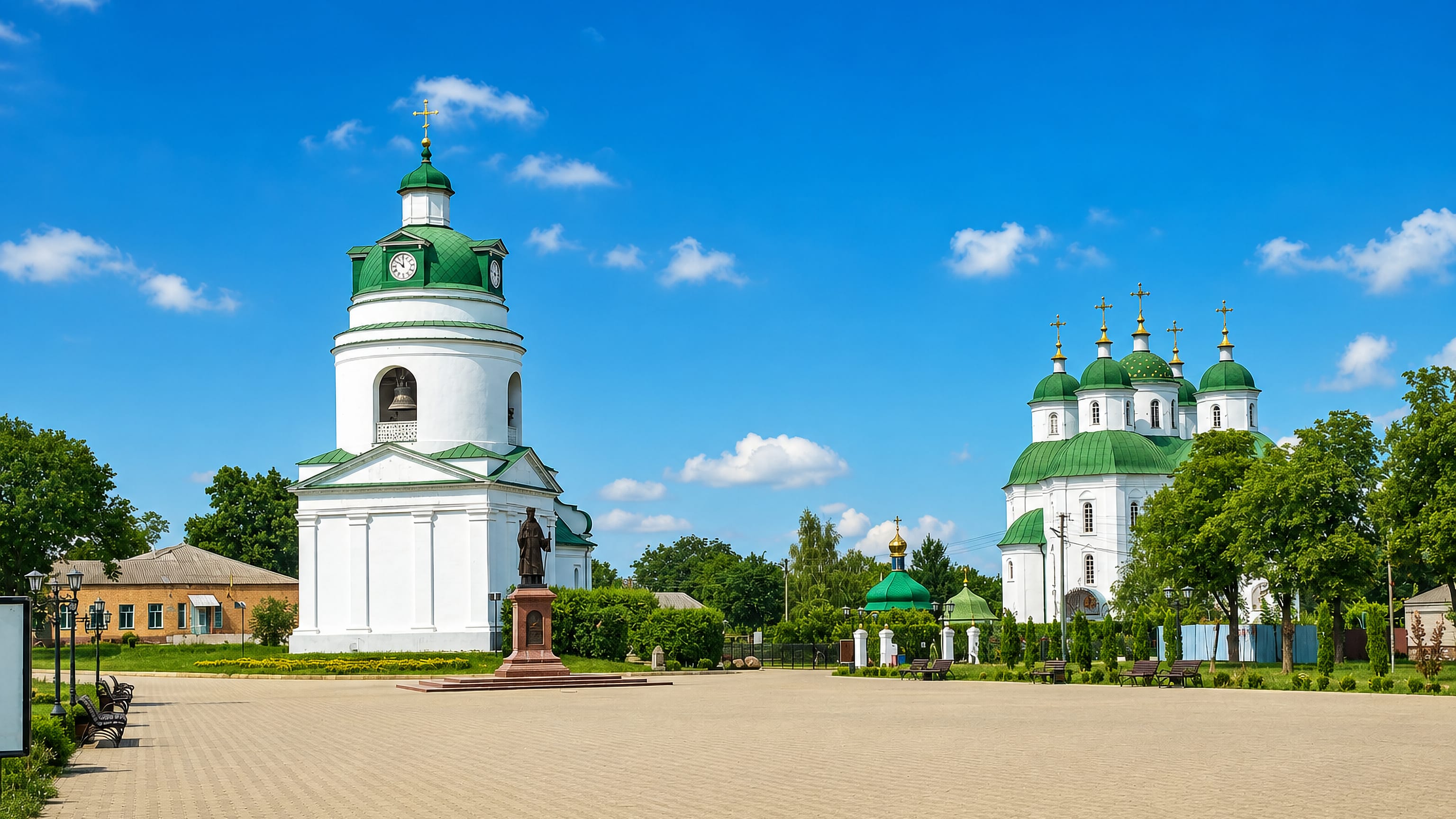 White-and-green Orthodox cathedral and free-standing bell tower on a paved central square in Pryluky, Ukraine.