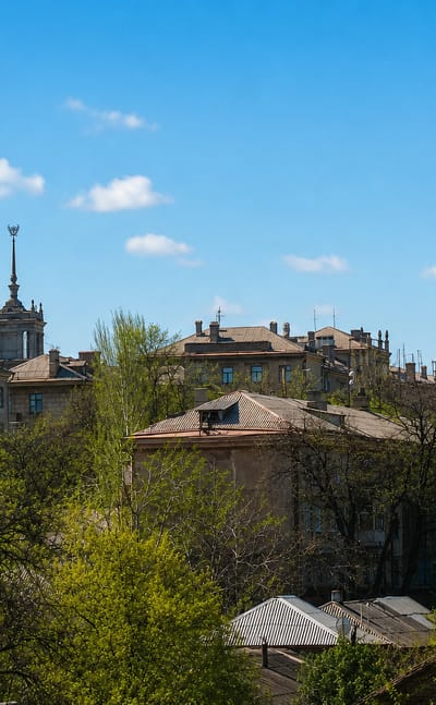 Rooftops of central Mariupol with two Stalinist spired towers rising above leafy trees under a blue sky.