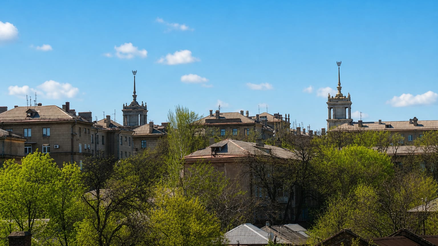 Rooftops of central Mariupol with two Stalinist spired towers rising above leafy trees under a blue sky.