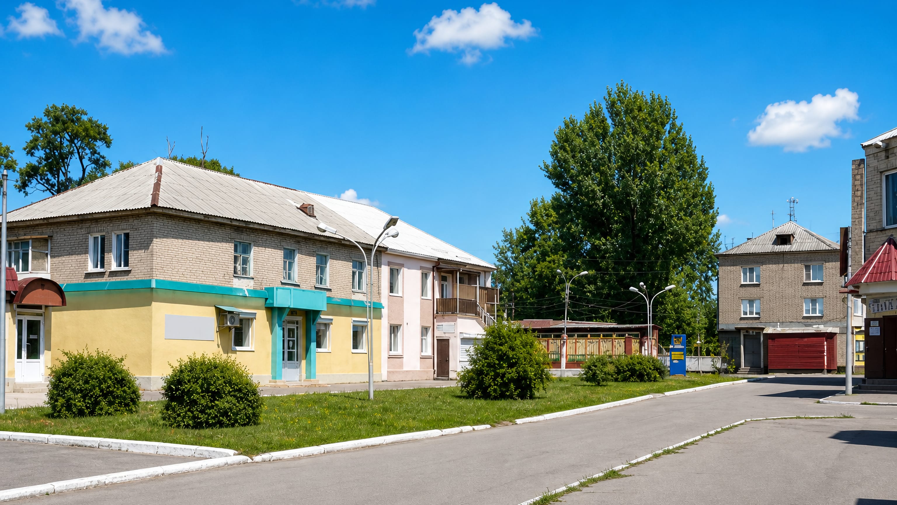Quiet sunlit street corner in Pokrovsk, Ukraine, with low brick and pastel-rendered buildings and roadside greenery.