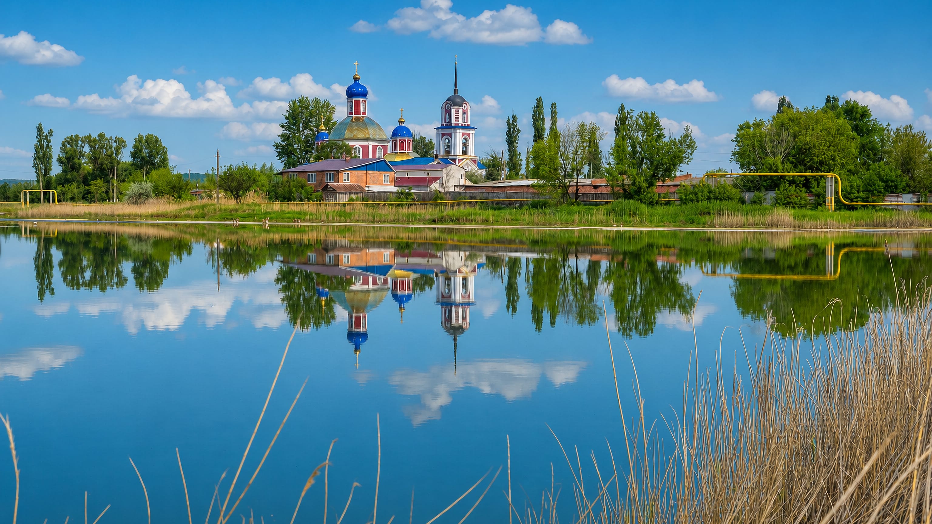 A blue-domed Orthodox church and bell tower mirrored in a calm lake on the rural edge of Slovyansk, Ukraine.