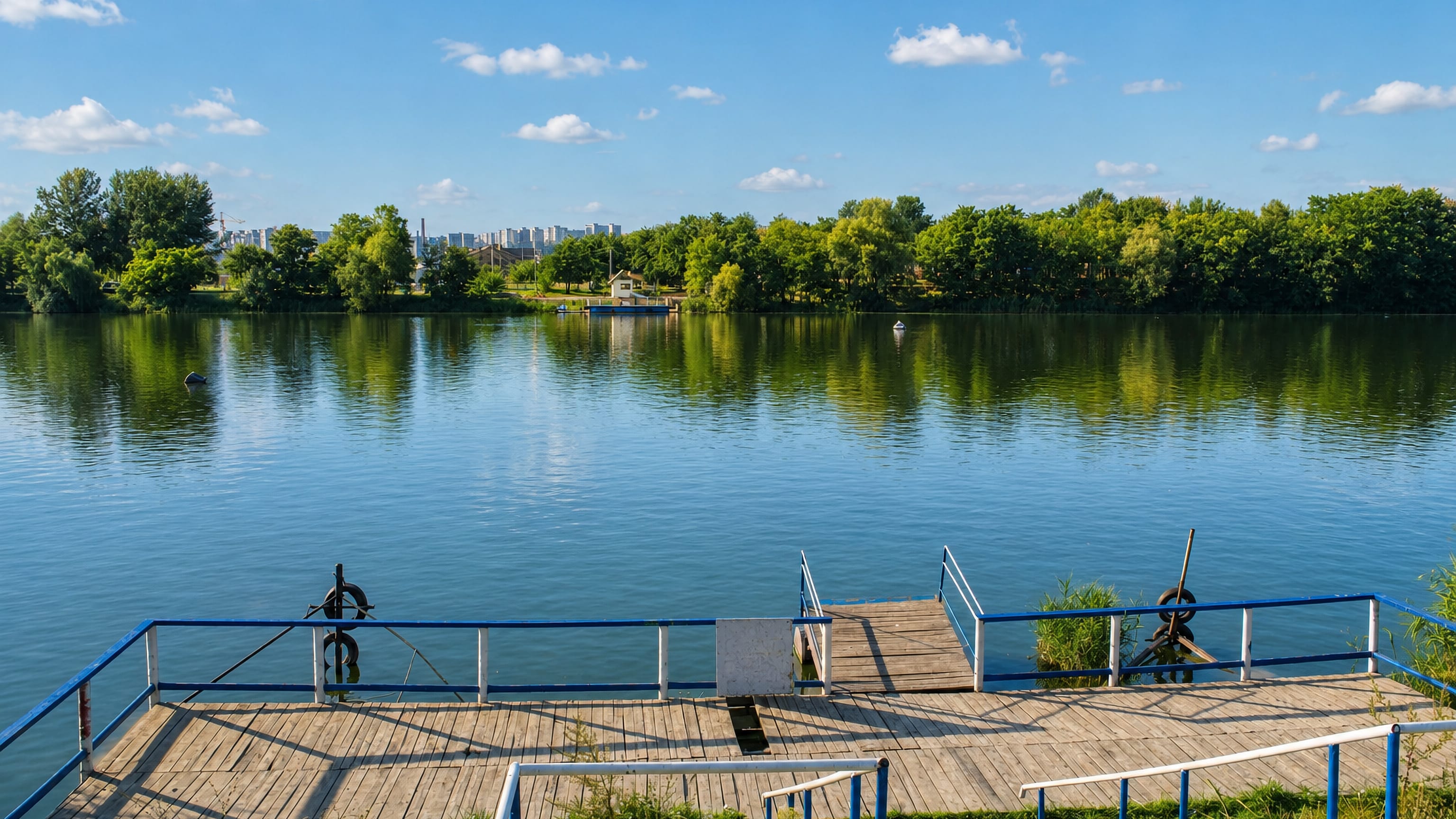 Wooden jetty on the tree-lined river at Zhuravlivka, Kharkiv, with distant apartment towers under a summer sky.