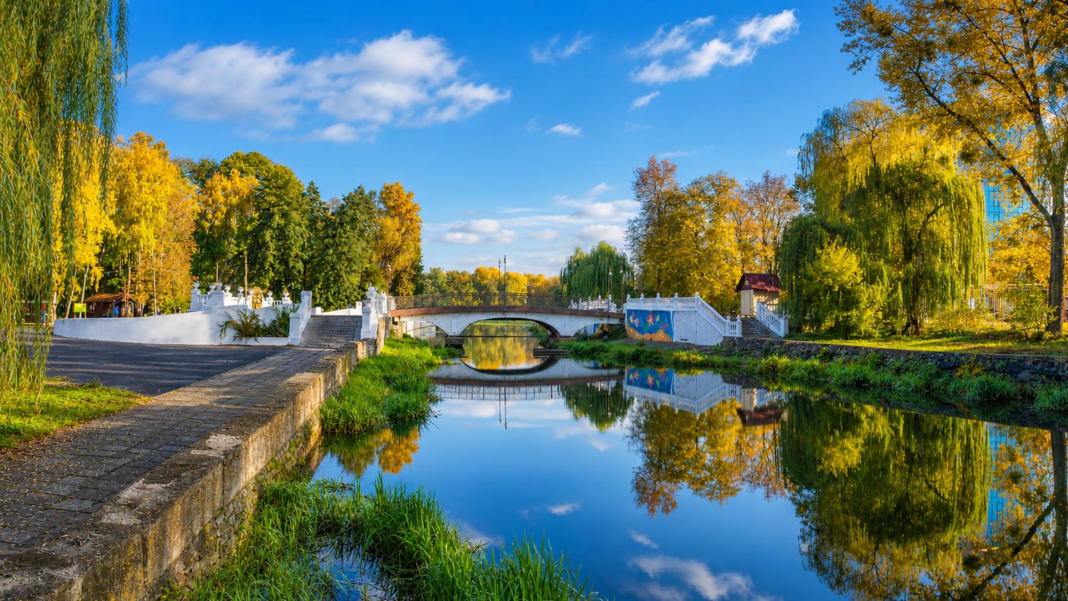 Autumn park scene in Khmelnytskyi, Ukraine, with a small stone bridge over a calm reflecting river lined with willows
