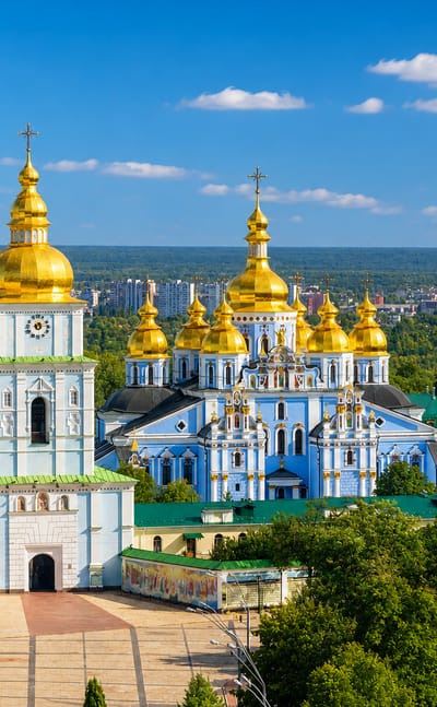 Kyiv's blue-and-gold St. Michael's Golden-Domed Monastery framed by green treetops under a clear sky.